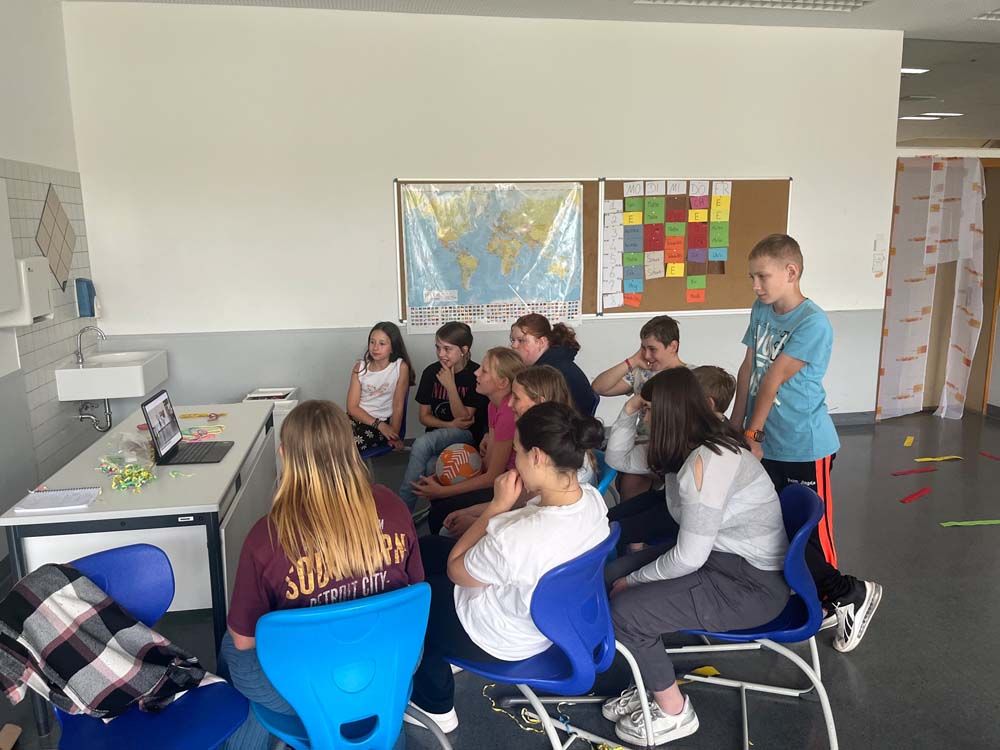 Eine Gruppe von Schülern schaut in einem Klassenzimmer auf einen Laptop-Bildschirm; an der Wand hängt eine Weltkarte.