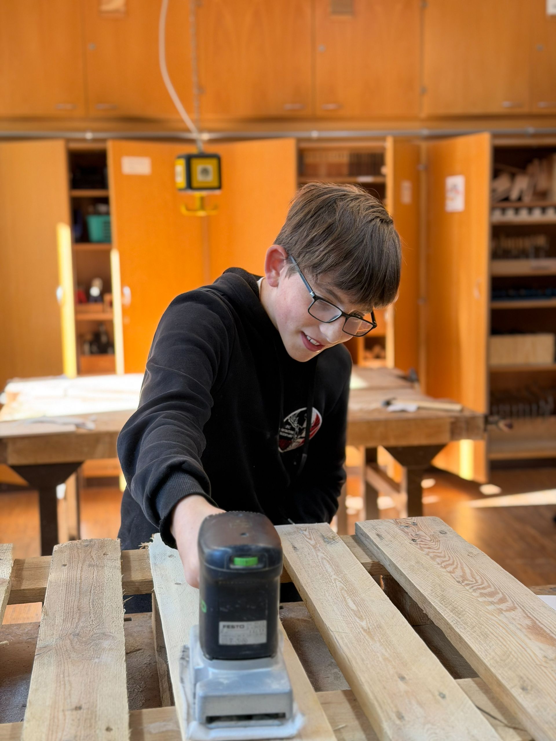Junger Mensch benutzt in einer Werkstattumgebung einen Schleifer an einer Holzpalette.