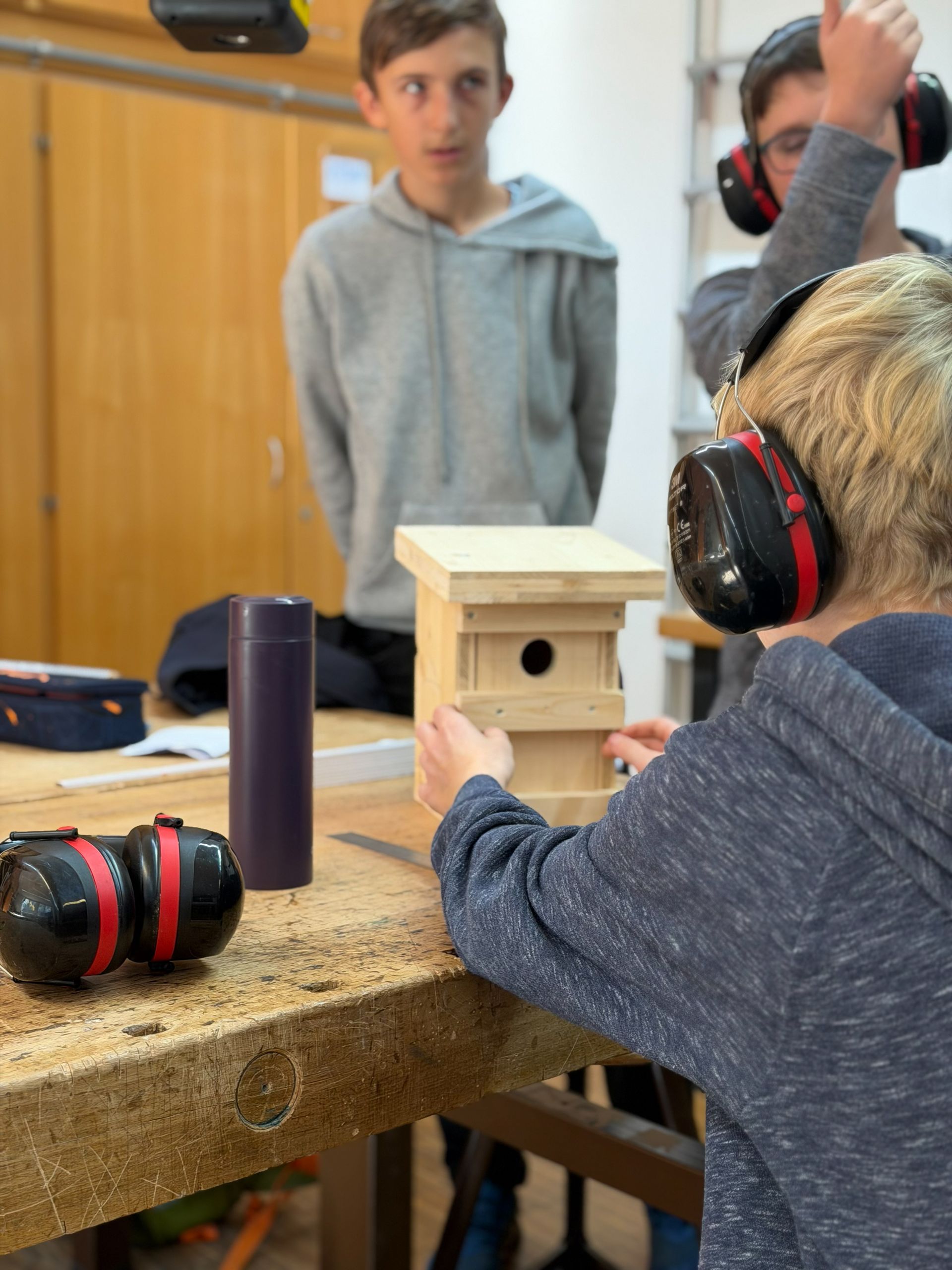 Jungen bauen in einer Werkstatt ein Vogelhaus und tragen dabei Gehörschutz; hölzerne Werkbank.