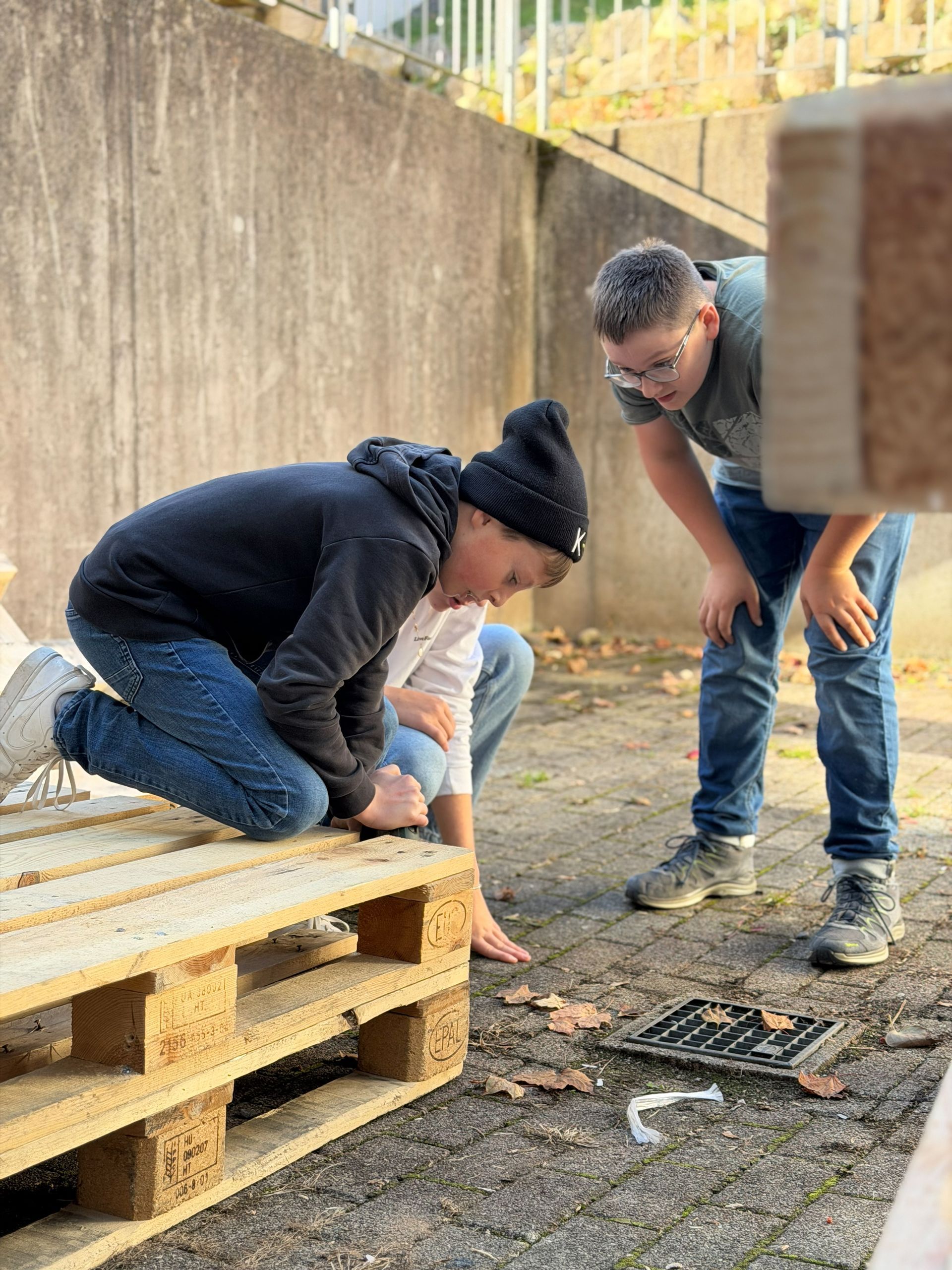 Zwei Personen untersuchen den Boden neben Holzpaletten; Außenaufnahme.