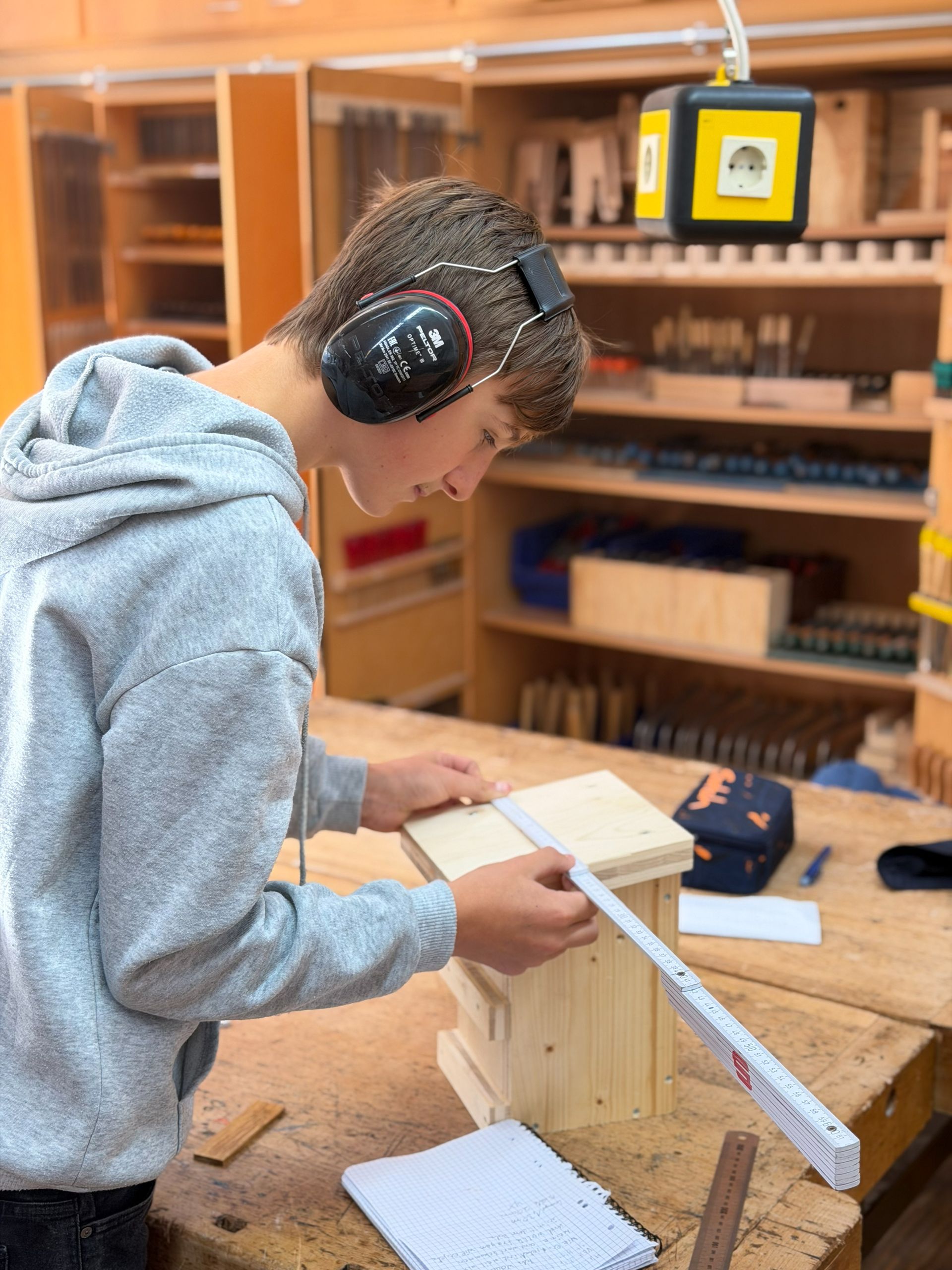 Eine Person, die in einer Werkstatt ein hölzernes Vogelhaus mit einem Lineal ausmisst. Sie trägt Ohrenschützer und konzentriert sich.