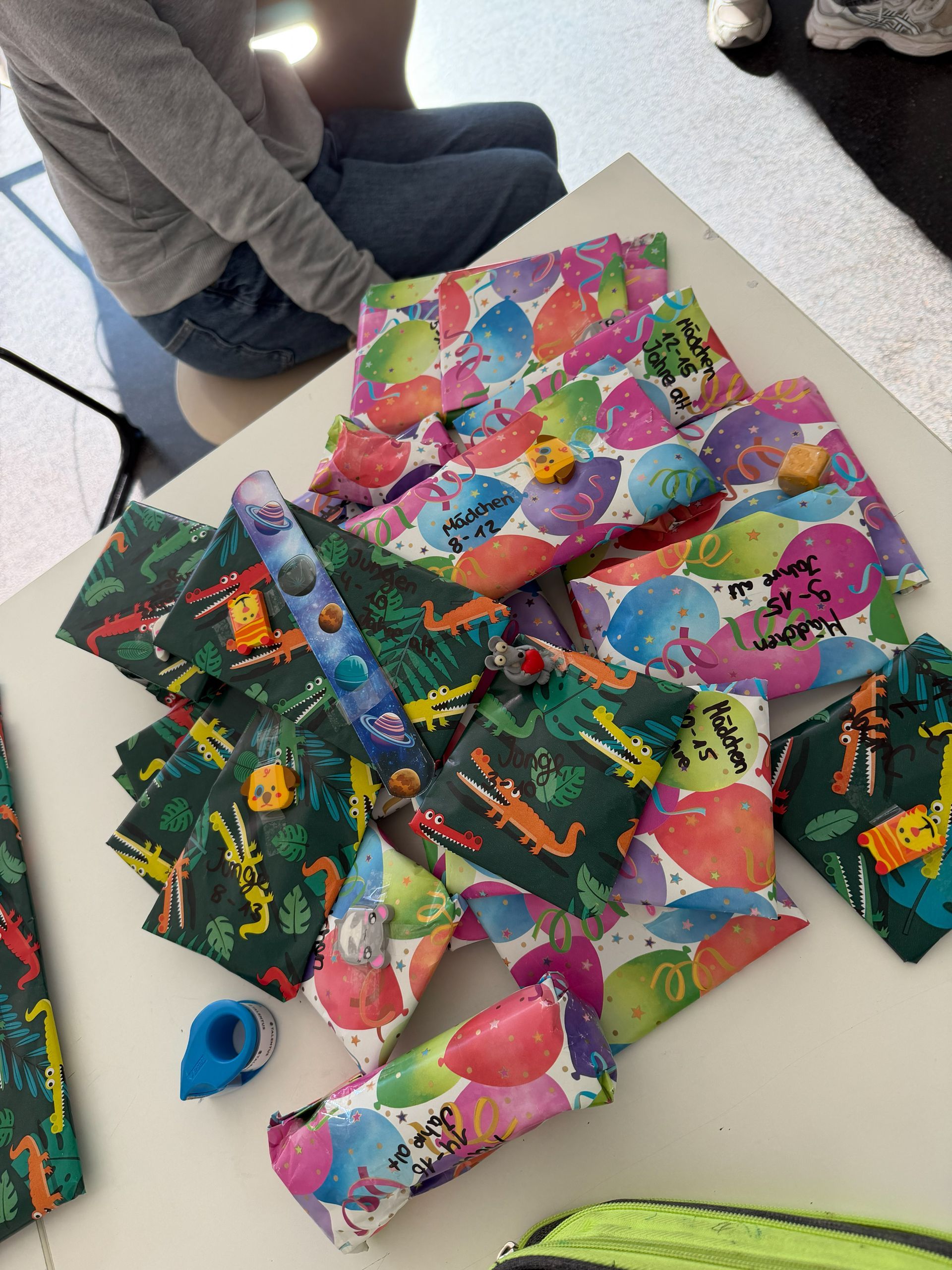 Ein Stapel bunter, verpackter Geschenke auf einem weißen Tisch. Jemand sitzt daneben.