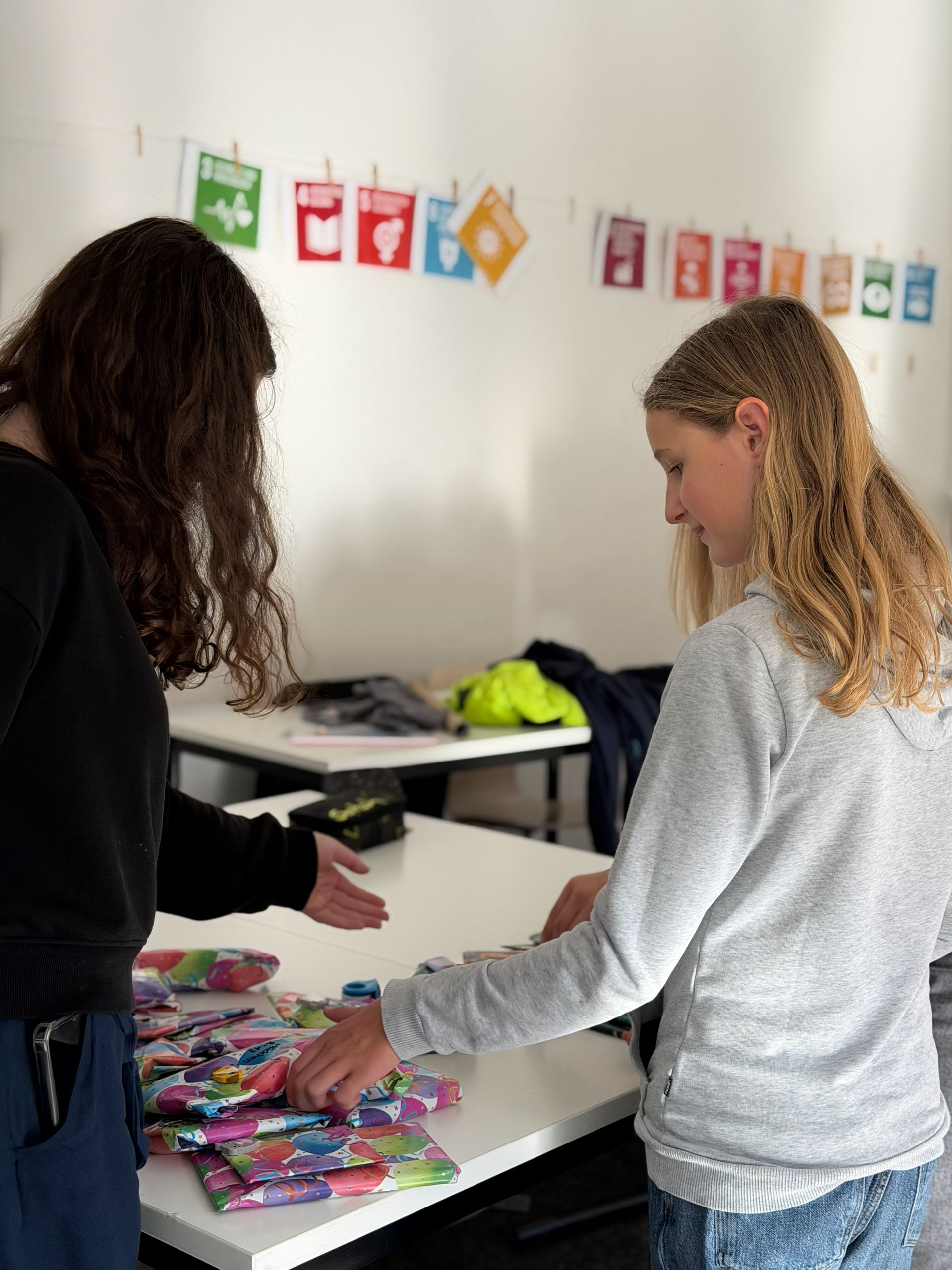 Zwei Personen verpacken Geschenke an einem Tisch. An der Wand hängen die UN-Nachhaltigkeitsziele.