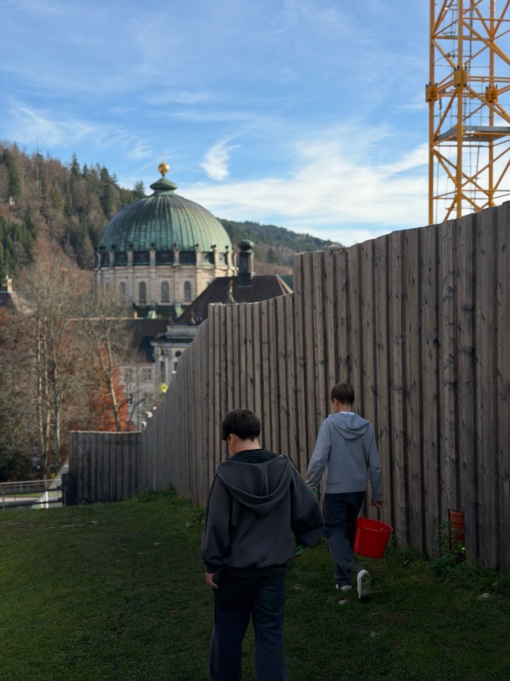 Zwei Personen gehen auf ein großes, kuppelförmiges Gebäude zu, vorbei an einem Holzzaun und einem Kran. Grünes Gras, blauer Himmel.