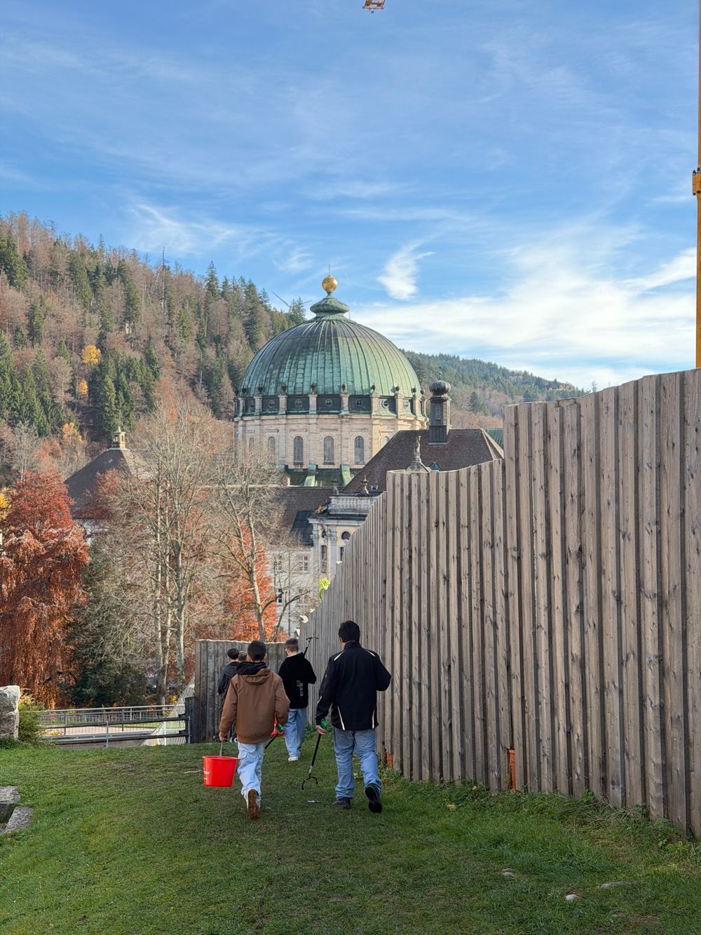 Drei Personen gehen an einem sonnigen Tag an einem Holzzaun vorbei auf ein kuppelförmiges Gebäude zu.