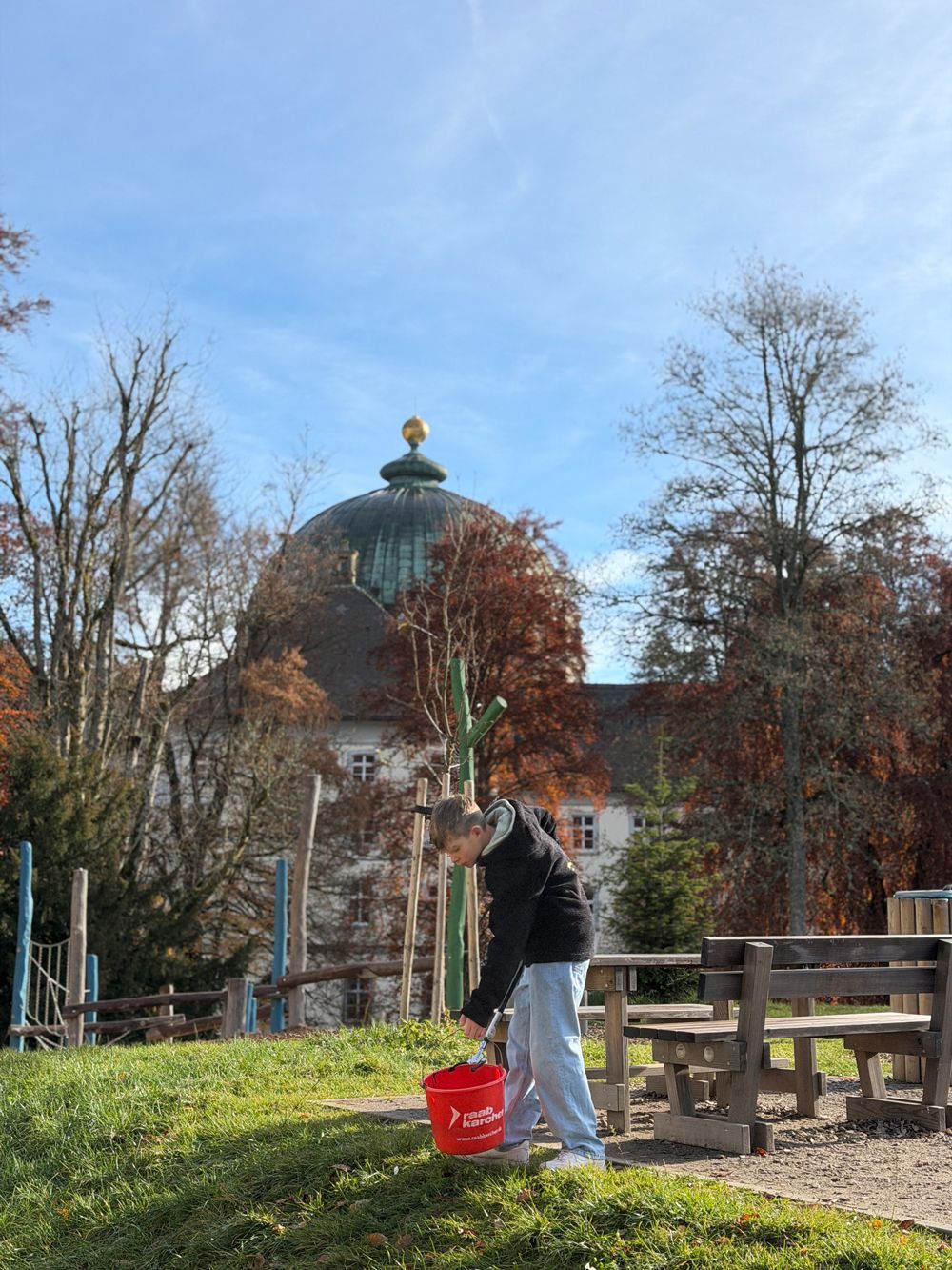 Eine Person hält einen roten Eimer in einem Park, im Hintergrund ein Gebäude mit Kuppel. Herbstliche Bäume, blauer Himmel.