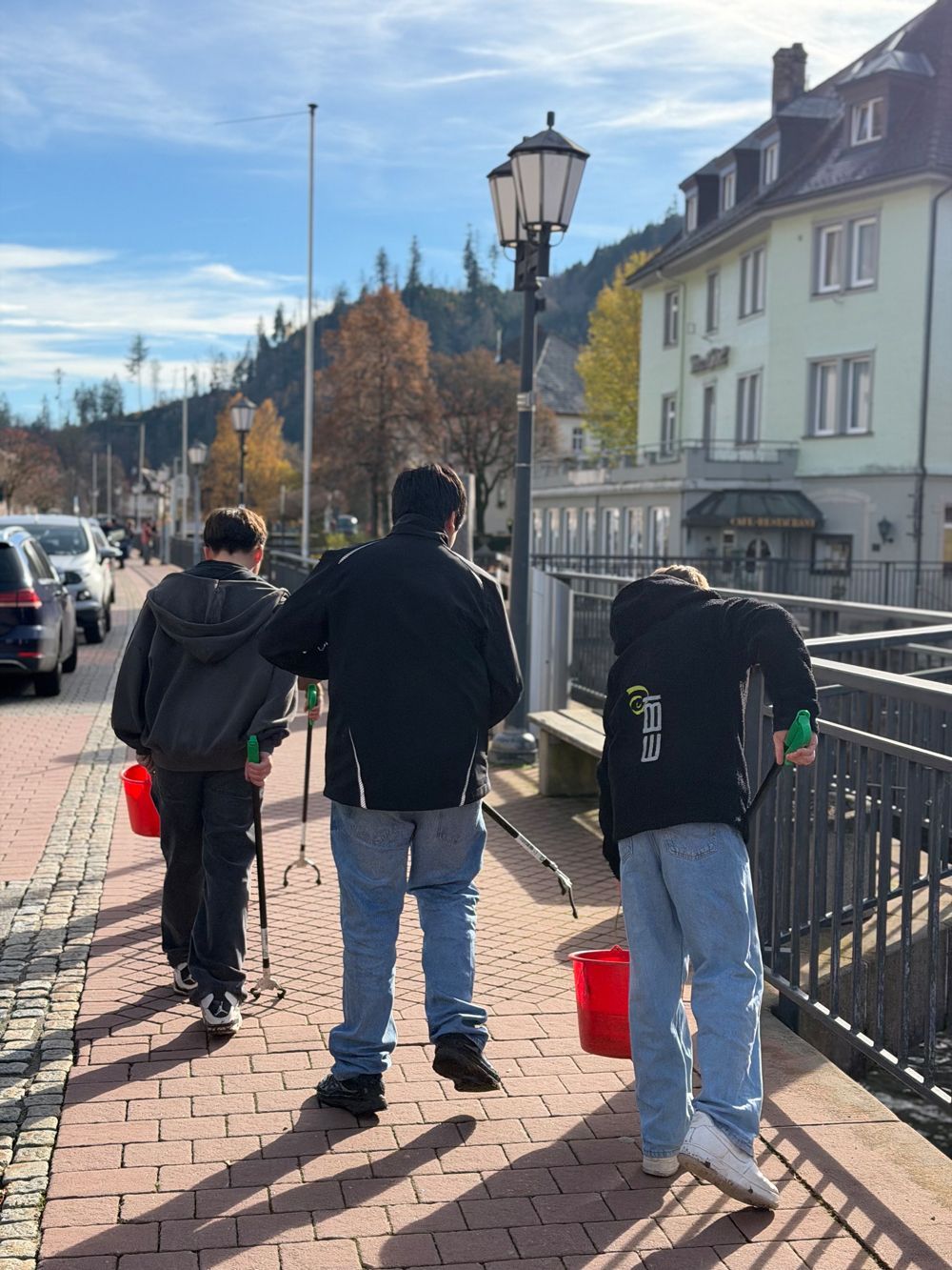 Drei Personen gehen mit Eimern und Stangen einen gepflasterten Weg entlang. Im Hintergrund sind Gebäude und Autos zu sehen.