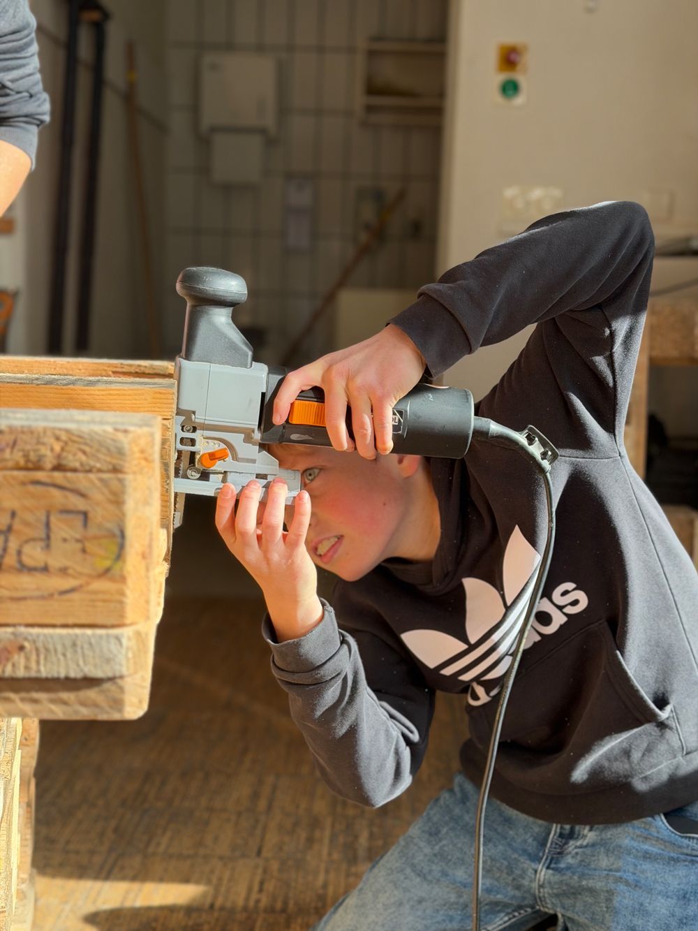Eine Person arbeitet mit einer Stichsäge an einer Holzpalette. Die Person trägt einen schwarzen Adidas-Hoodie und ist konzentriert bei der Arbeit.