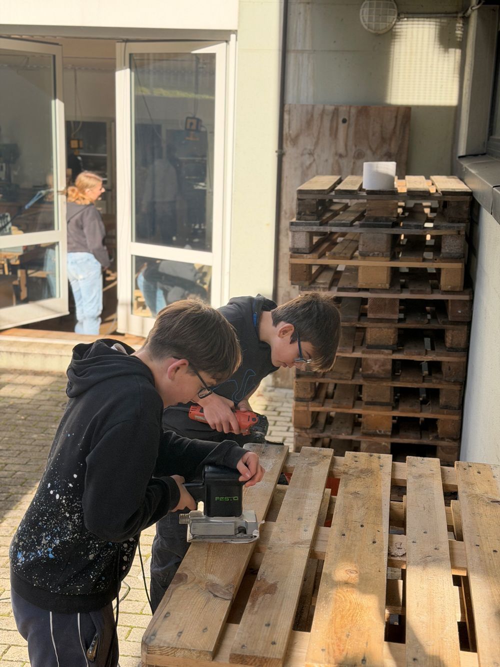 Zwei Personen arbeiten draußen an Holzpaletten mit Elektrowerkzeugen; eine Person ist im Türrahmen zu sehen.