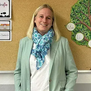 Frau im Blazer und mit Schal, lächelnd, steht vor einer Pinnwand mit Unterrichtsmaterialien.