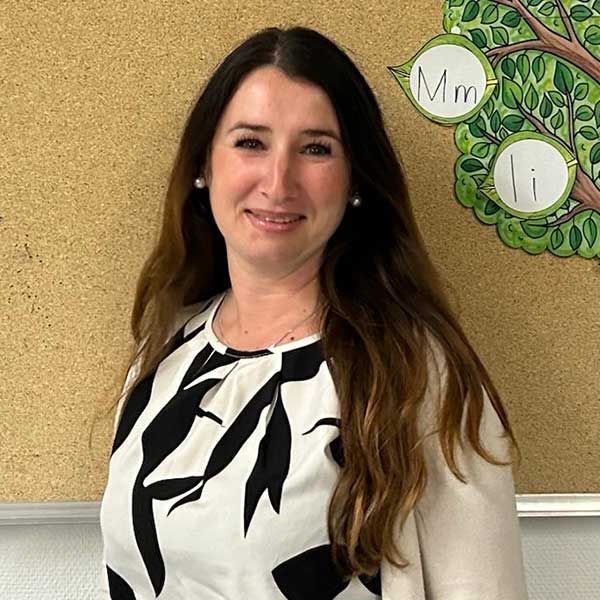 Eine Frau mit langen braunen Haaren, die ein schwarz-weiß gemustertes Hemd trägt, lächelt in einem Klassenzimmer, an dessen Wand ein Baumdiagramm hängt.