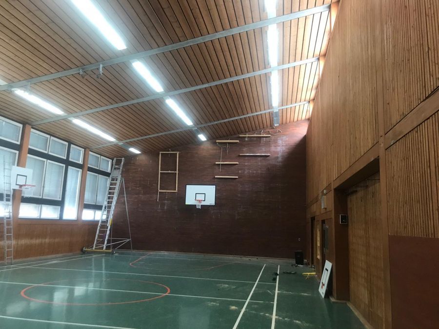 Turnhalleninterieur mit Basketballkörben, grünem Boden, holzvertäfelten Wänden und heller Deckenbeleuchtung.