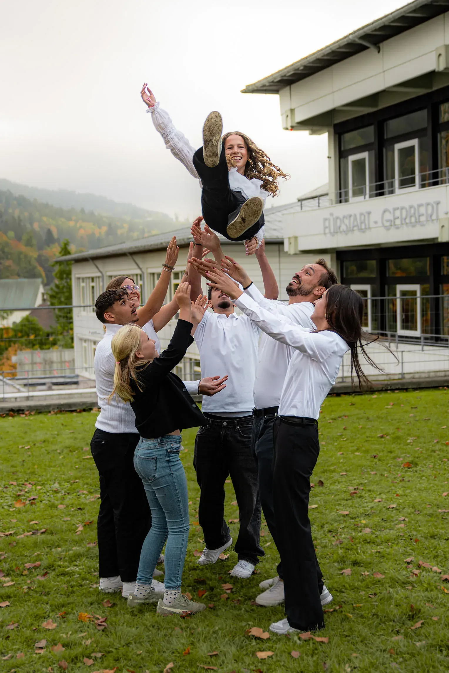 Eine Gruppe von Menschen wirft eine Person auf einem Rasen vor einem Gebäude in die Luft.