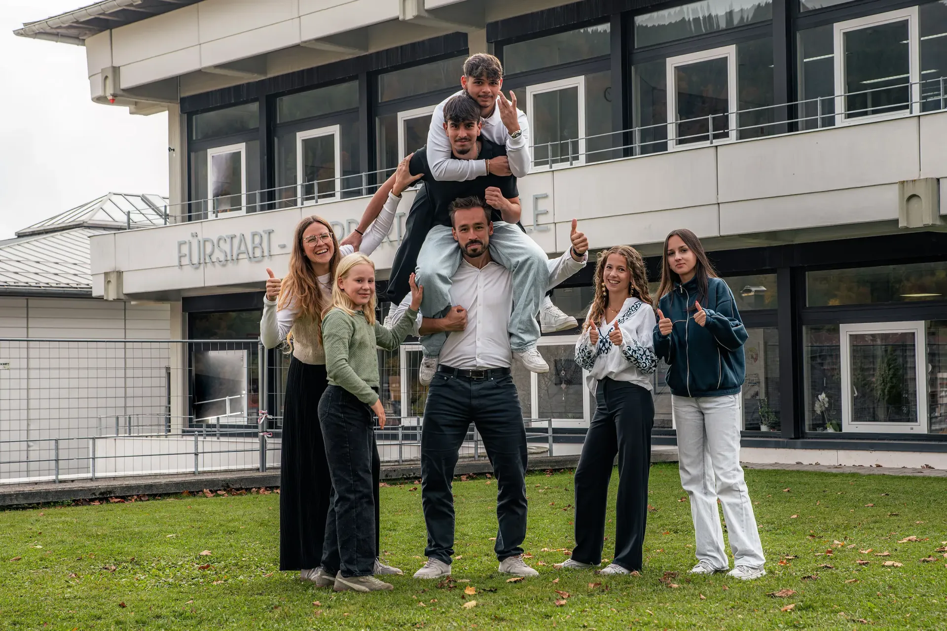 Eine Gruppe von Menschen posiert vor einem Gebäude. Einige zeigen den Daumen nach oben, manche stehen auf anderen.