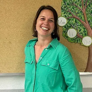 Eine Frau in einem grünen Hemd lächelt vor einer Pinnwand mit einem ausgeschnittenen Baum und Buchstaben des Alphabets.