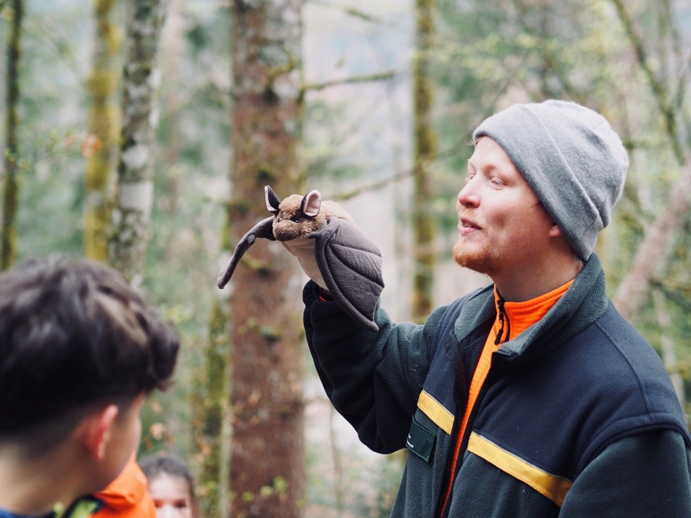 Eine Person mit grauer Mütze, die in einem Wald einen Schläger hochhält und zu einer kleinen Gruppe spricht