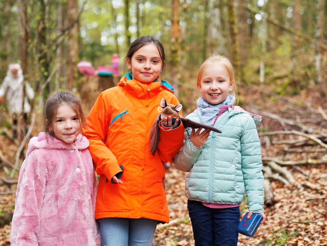 Drei Kinder im Freien in einem Wald, eines davon in einer orangefarbenen Jacke, das einen Vogel hält.