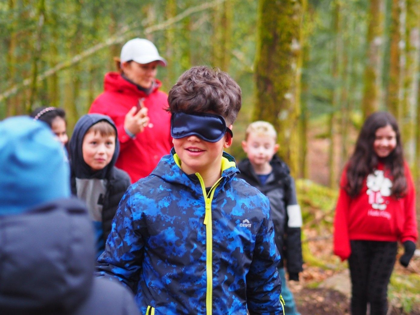 Ein Kind mit verbundenen Augen und blauer Jacke steht mit einer Gruppe von Menschen auf einer Waldlichtung.