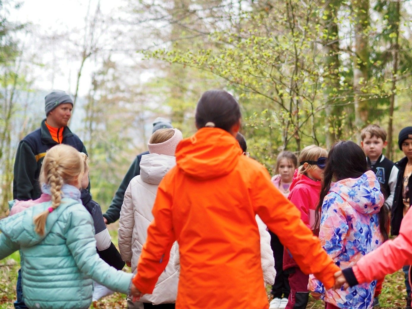 Kinder in bunten Jacken halten sich im Freien an den Händen und bilden einen Kreis; ein Erwachsener ist in der Nähe.
