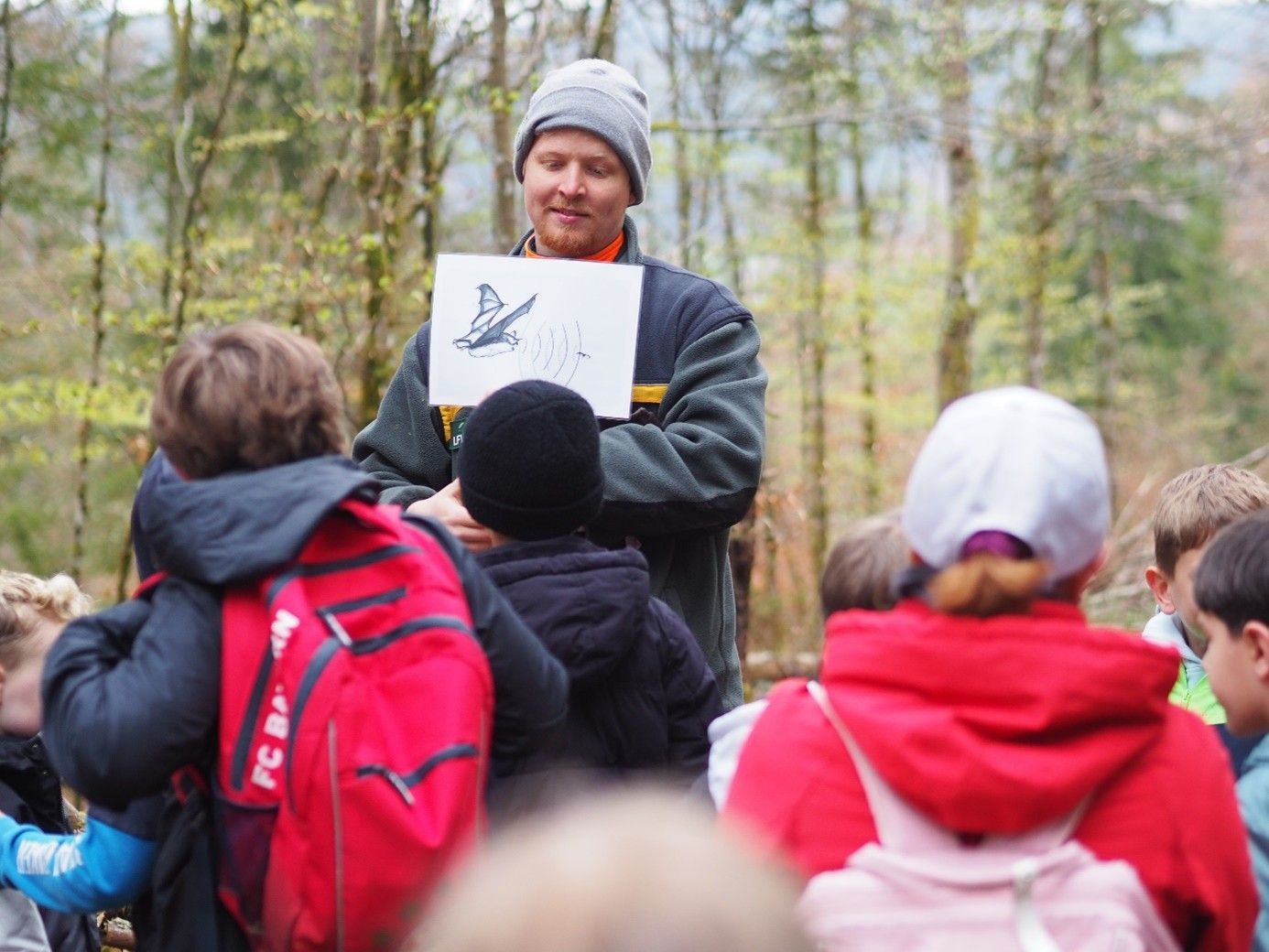 Eine Lehrerin hält eine Zeichnung in der Hand, während sie im Freien in einem Waldgebiet mit einer Gruppe Kinder spricht.
