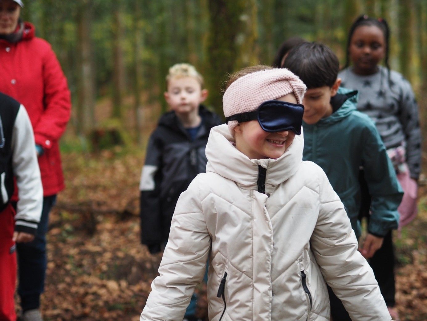 Kinder im Freien in einem Wald, eines trägt eine Augenbinde und eine Winterjacke, die anderen stehen hinter ihr.