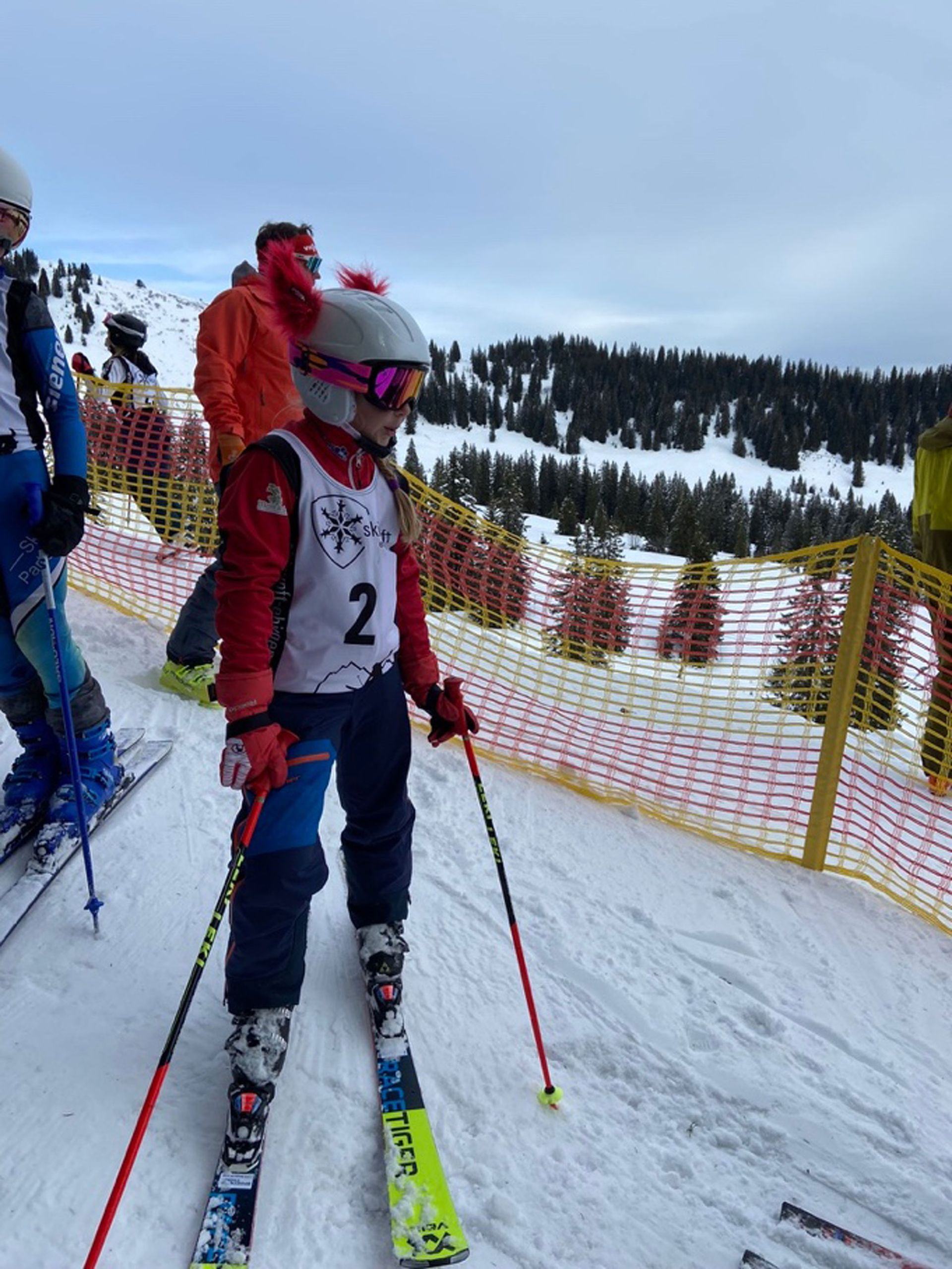 Ein Skifahrer mit der Startnummer 2 steht mit den Skistöcken in der Hand auf dem Gipfel eines schneebedeckten Berges und bereitet sich auf ein Rennen vor.