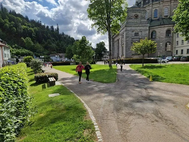 Zwei Personen rennen auf einem Weg in einem Park, in der Nähe eines Gebäudes. Bäume und grünes Gras umgeben sie.