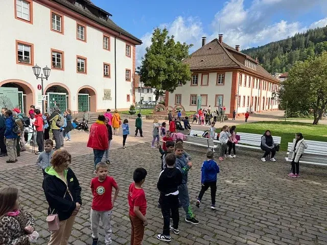 Kinder und Erwachsene auf einem Marktplatz. Gebäude, Bänke und ein Baum sind zu sehen. Sonniger Tag.