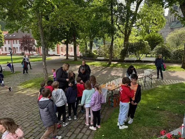 Kinder versammelten sich im Freien mit Erwachsenen; ein Tisch war gedeckt, im Hintergrund waren Bäume und Gebäude zu sehen.