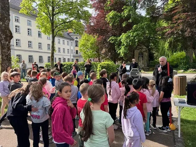 Eine Gruppe Kinder hört im Freien, in der Nähe eines Gebäudes und von Bäumen, einer Person mit Mikrofon zu.