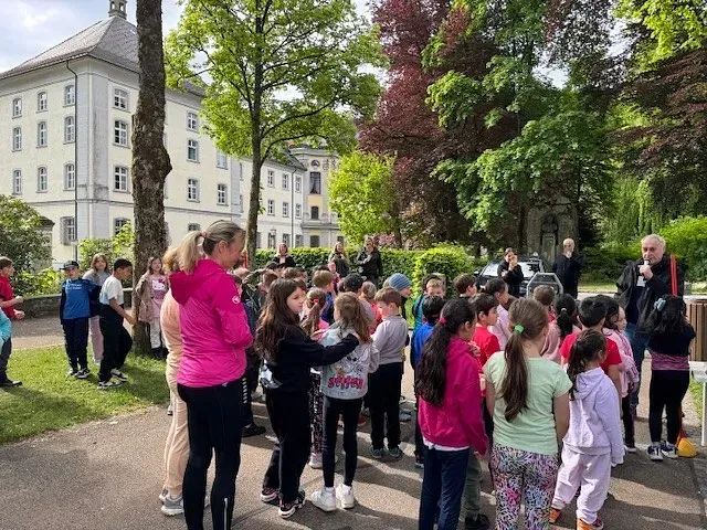 Gruppe von Kindern im Freien mit Erwachsenen; im Hintergrund mehrere Gebäude.