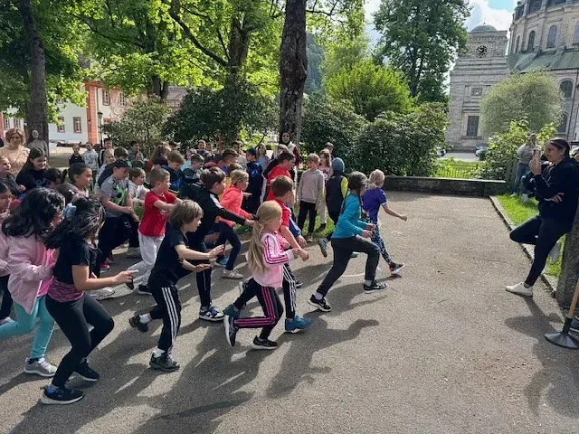 Kinder treiben Sport im Freien und folgen dabei einem Trainer. Im Hintergrund sind grüne Bäume und ein Gebäude zu sehen.