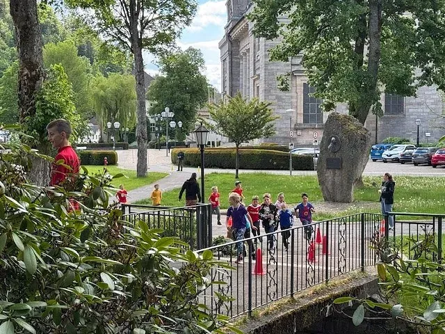 Kinder rennen über eine Brücke in einem Park, im Hintergrund eine Kirche, Bäume und Grünanlagen.