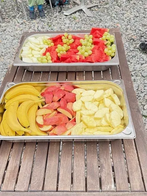 Zwei Metalltabletts mit geschnittenem Obst: Bananen, Äpfel, Wassermelone und Weintrauben auf einem Holztisch.
