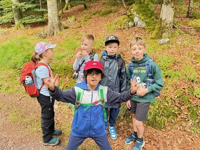 Fünf Kinder posieren in einem Wald. Eines im Vordergrund zeigt das Friedenszeichen, die anderen lächeln. Bäume und Sträucher umgeben sie.