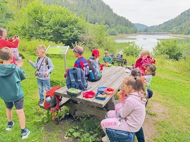 Kinder sitzen an einem Picknicktisch an einem See und essen zu Mittag. Einige schauen etwas an.