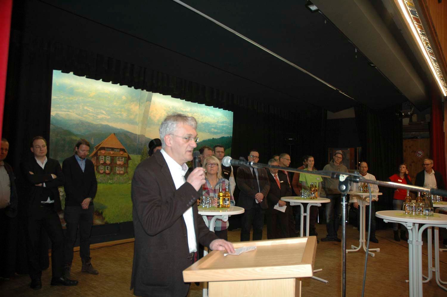 Ein Mann spricht an einem Rednerpult zu einer Gruppe in einem Saal vor einer Landschaftskulisse.