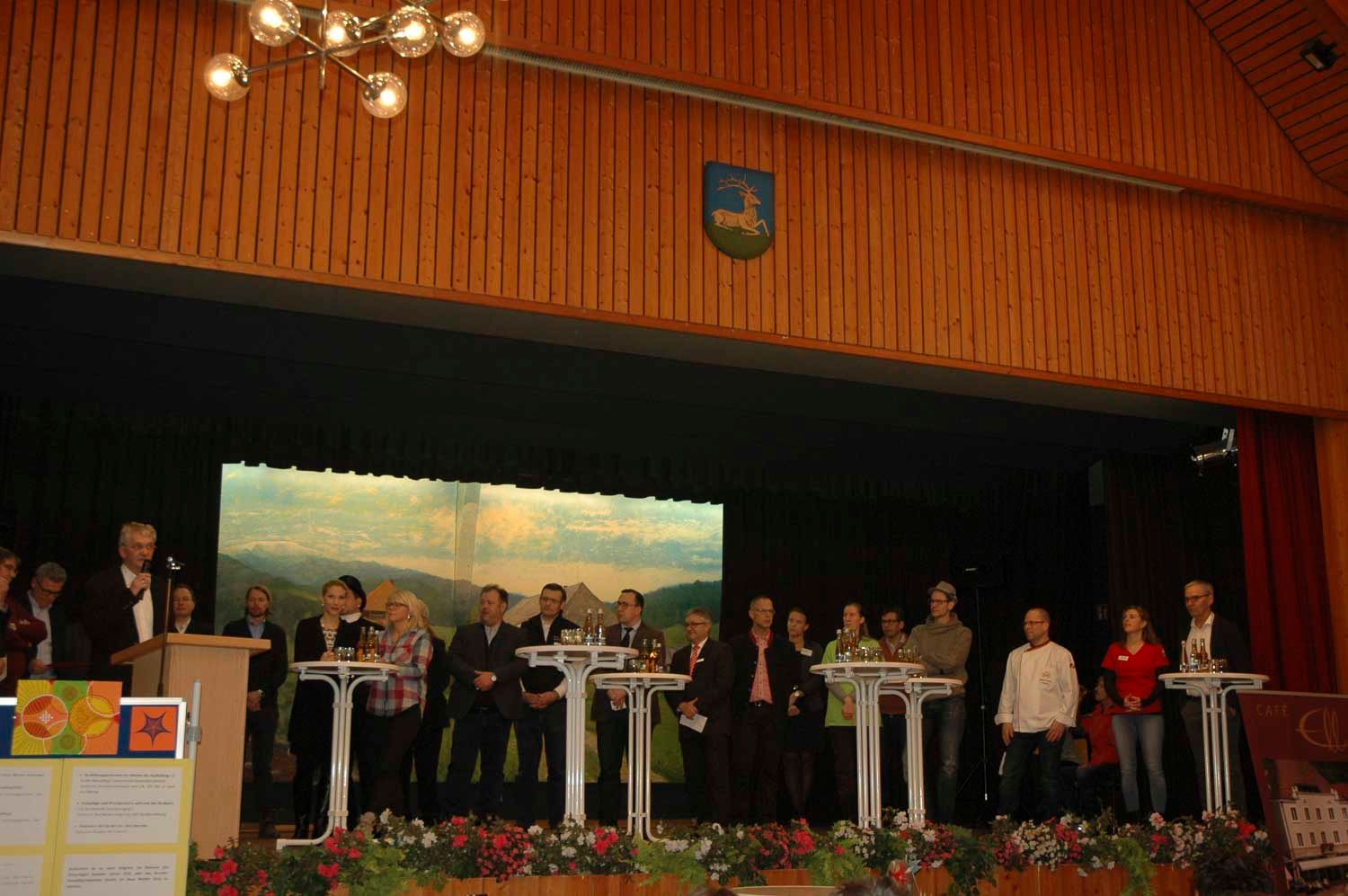 Menschen stehen auf einer Bühne mit Rednerpulten, vermutlich bei einer politischen Veranstaltung. Im Hintergrund ist eine große Leinwand zu sehen.
