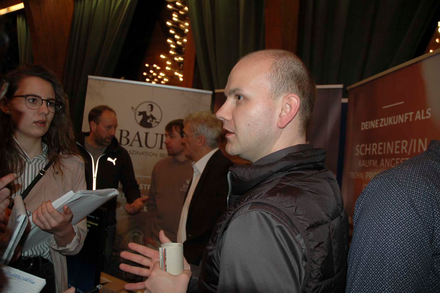 Ein Mann in dunkler Weste spricht, umringt von Menschen, im Hintergrund sind Banner zu sehen.