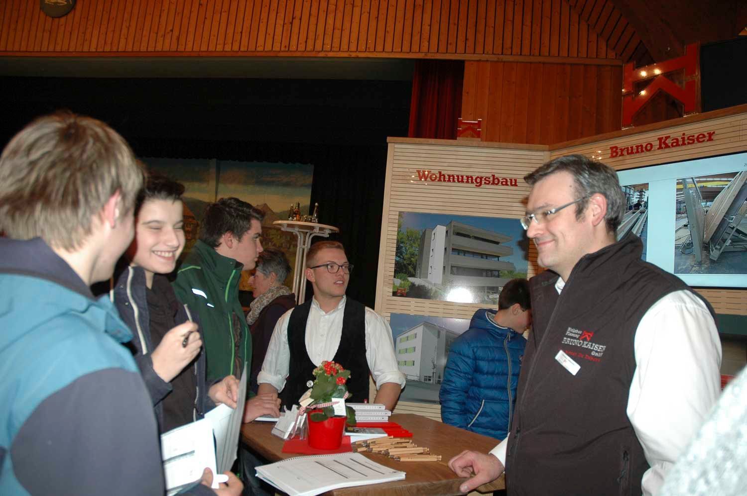 Menschen an einem Messestand, unterhalten sich und lächeln. Ein Schild verkündet „Wohnungsbau“.