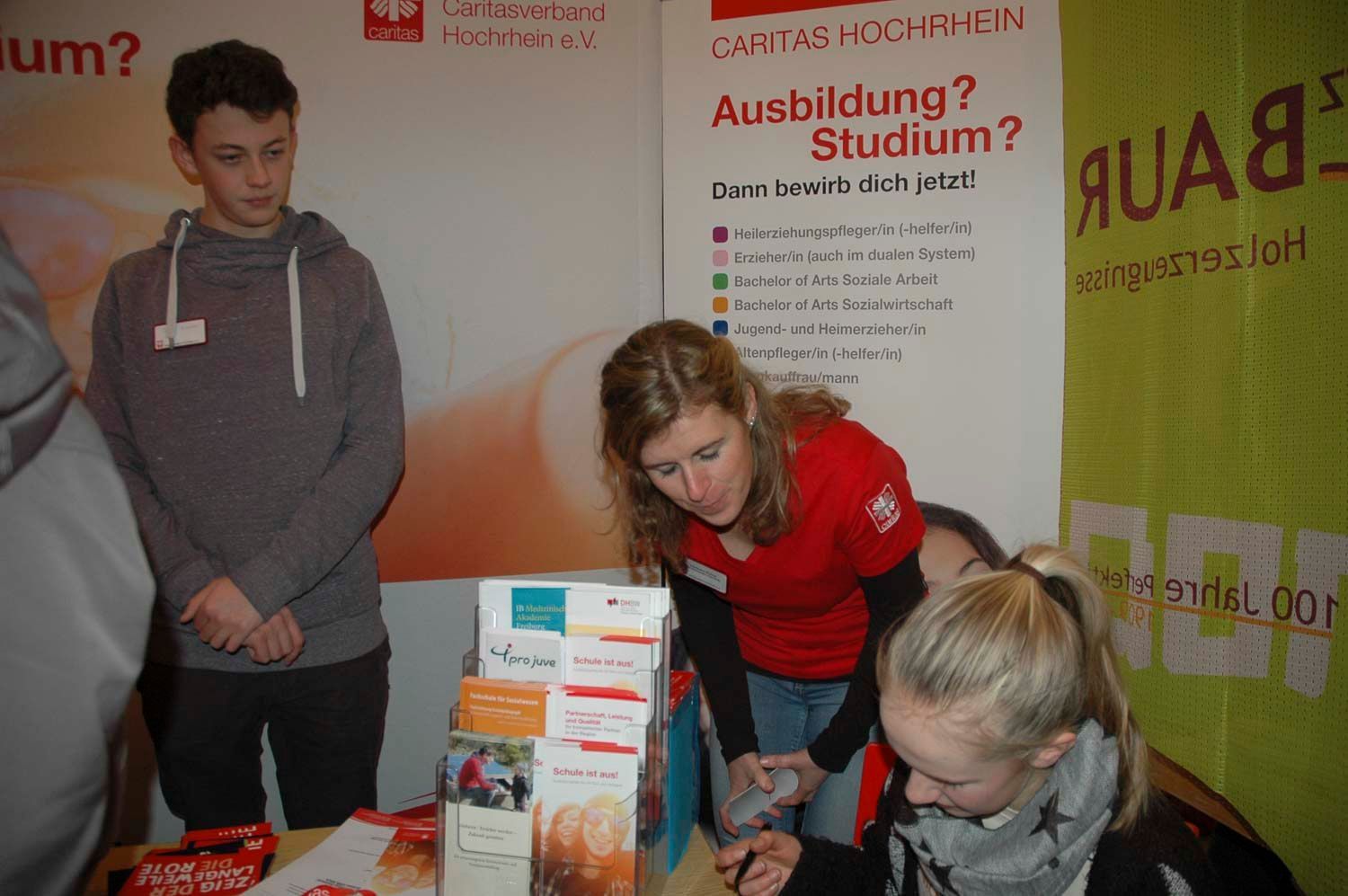 Menschen an einem Informationsstand. Eine Frau im roten Hemd hilft einer Schülerin; eine andere schaut zu. Plakate sind zu sehen.