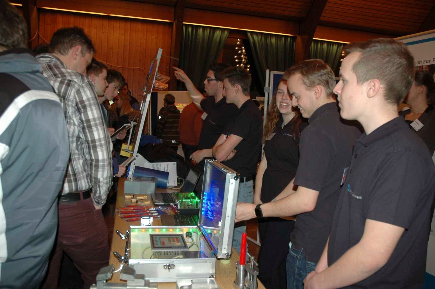 Menschen versammeln sich an einem Technikstand und betrachten Geräte und einen Computer. Einige tragen schwarze Hemden.