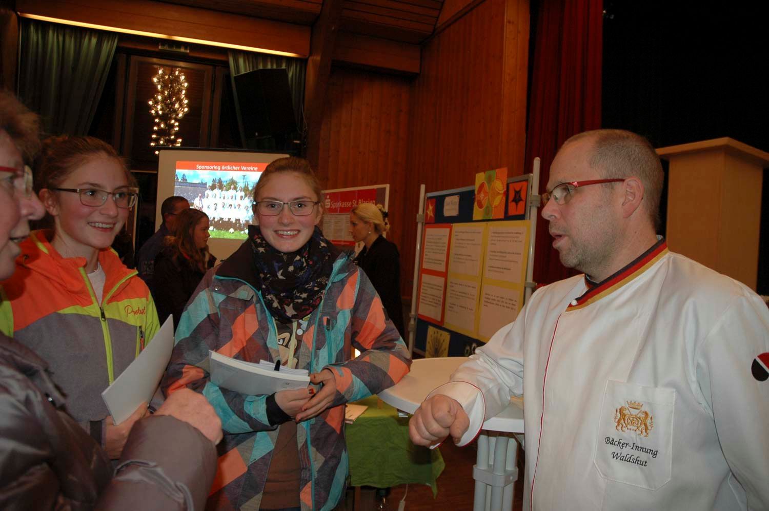Ein Koch unterhält sich mit zwei lächelnden Mädchen, möglicherweise auf einer Veranstaltung. Die Mädchen tragen weiße Kochjacken und halten Papiere in den Händen.