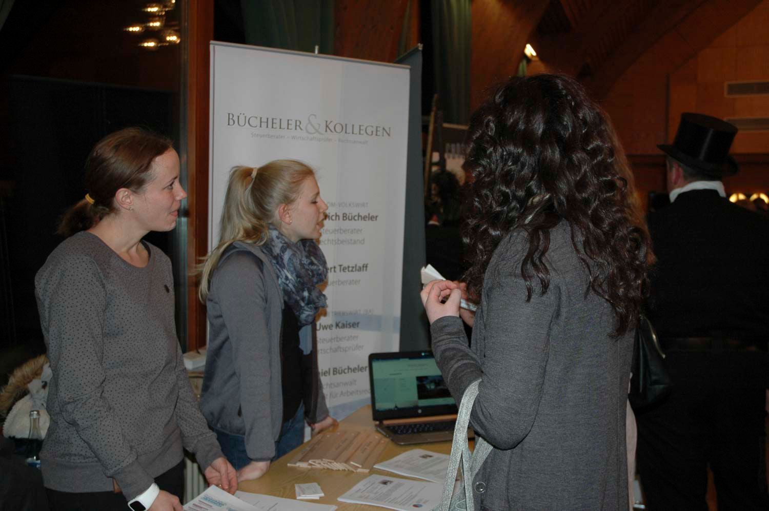 Drei Frauen an einem Stand mit einem Banner. Eine Frau spricht, die anderen hören zu. Schreibtisch mit Laptop und Unterlagen.