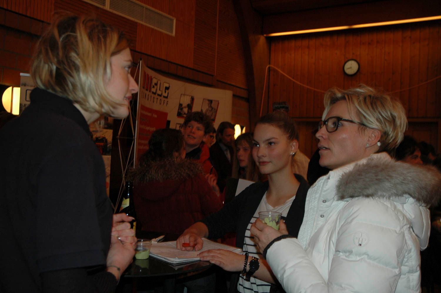 Bei einer Veranstaltung unterhalten sich zwei Personen; eine trägt eine weiße Jacke mit Pelzkapuze, die andere eine dunkle Jacke. Hintergrund: Holzvertäfelung.