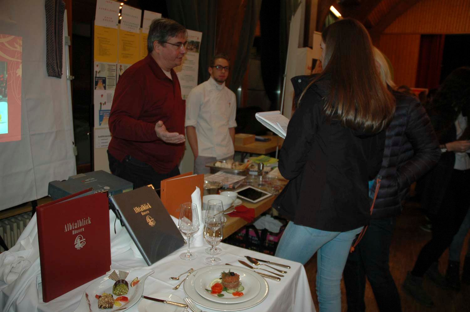 Ein Mann deutet auf eine Auslage; ein Tisch mit Speisen, Speisekarten und Personen im Raum.