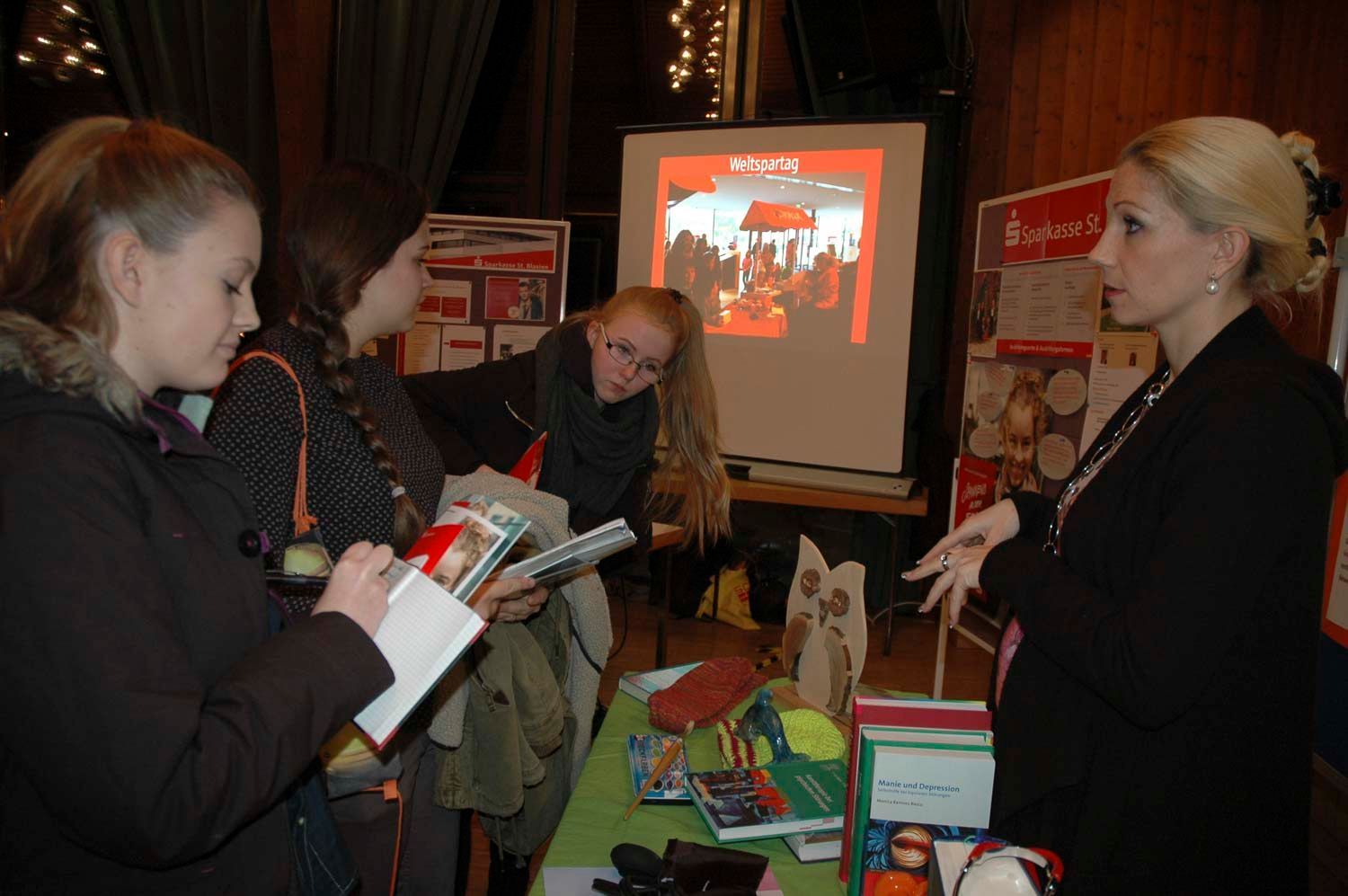 Menschen sitzen an einem Tisch mit Broschüren und einer Leinwand. Eine Frau spricht, drei andere betrachten die Gegenstände.