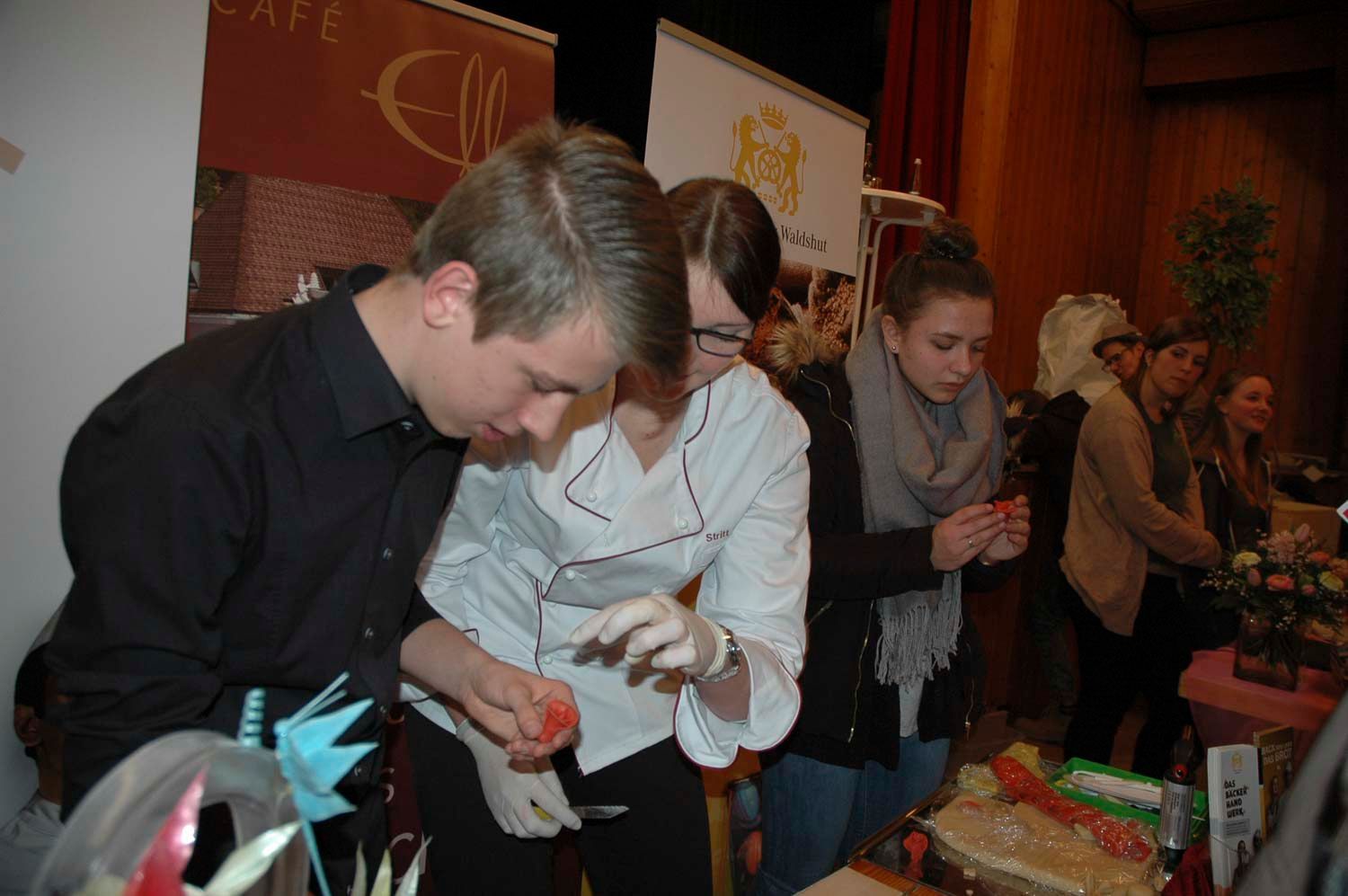 Menschen arbeiten an einem Auslagetisch mit Lebensmitteln. Zwei sind konzentriert bei der Sache; die anderen schauen zu.