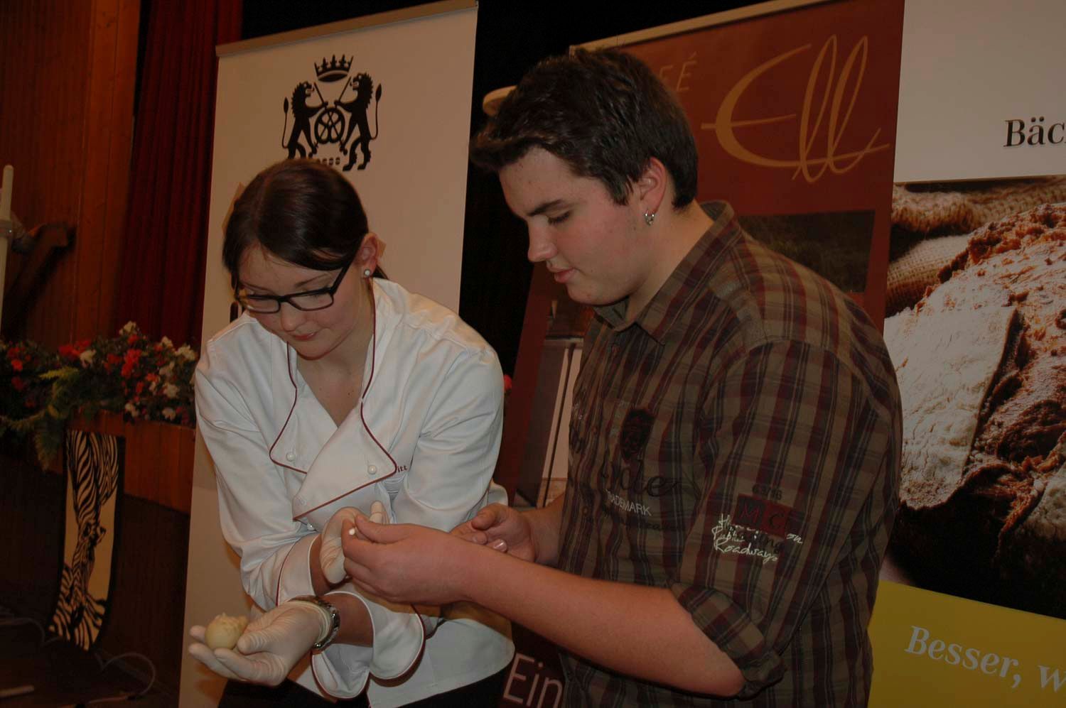 Zwei Personen bereiten drinnen Essen zu; eine Frau im Kochkittel, ein Mann im karierten Hemd, der Teig knetet.