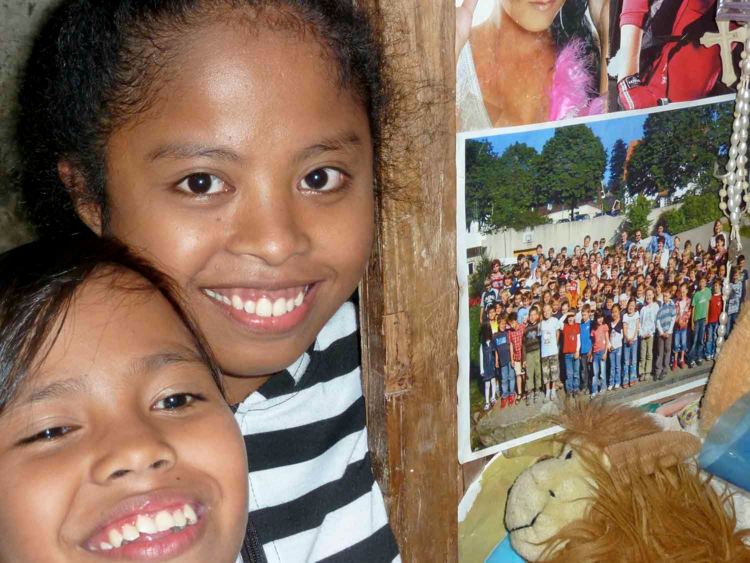 Zwei lächelnde Mädchen, eines davon mit einem gestreiften Hemd, posieren eng beieinander. Im Hintergrund sind Fotos zu sehen.